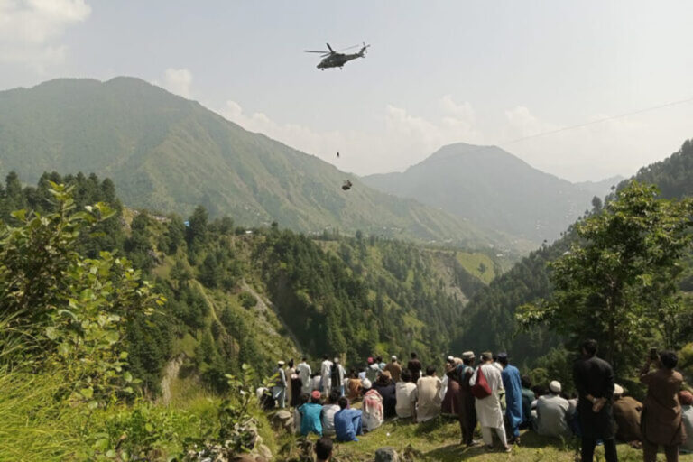 People watch as an army soldier slings down from a helicopter during a rescue mission to recover students stuck in a chairlift in Pashto village of mountainous Khyber Pakhtunkhwa province, on August 22, 2023. Six children and two adults were suspended inside a cable car dangling over a deep valley in Pakistan for several hours on August 22, as a military helicopter hovered nearby.
