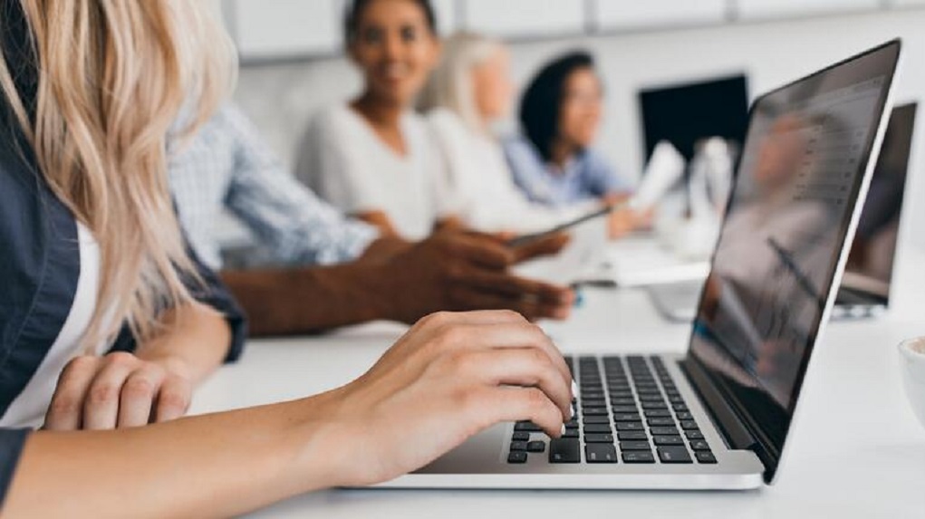 Blonde woman with elegant hairstyle typing text on keyboard in office. Indoor portrait of international employees with secretary using laptop on foreground..